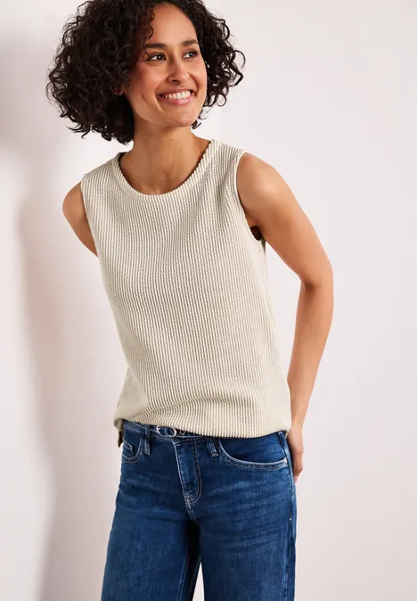 Persoon voor een witte achtergrond, gekleed in een mouwloze beige top en een blauwe spijkerbroek.