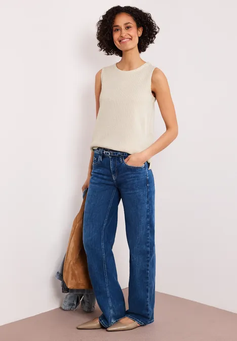 Vrouw in crèmetop en wijde spijkerbroek poseert nonchalant voor een muur.