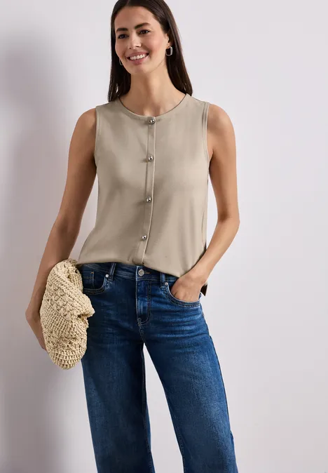 Eine Frau trägt eine ärmellose beige Bluse und blaue Jeans, hält eine geflochtene Tasche.