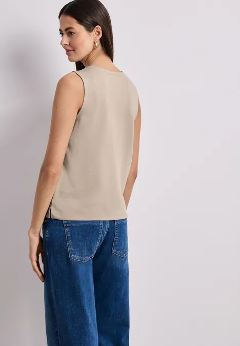 Frau steht mit dem Rücken zur Kamera, trägt ein beiges ärmelloses Top und weite blaue Jeans.