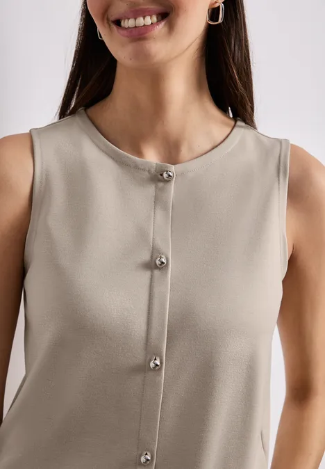 Eine Frau trägt ein ärmelloses, beiges Oberteil mit silbernen Knöpfen und lächelt.