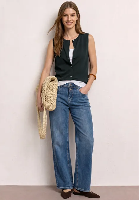 Weste aus Jersey mit Rundhals deep bound green Frau in weiten Jeans, schwarzem ärmellosem Top, weißem Shirt, Loafern, mit geflochtener Tasche vor hellem Hintergrund.