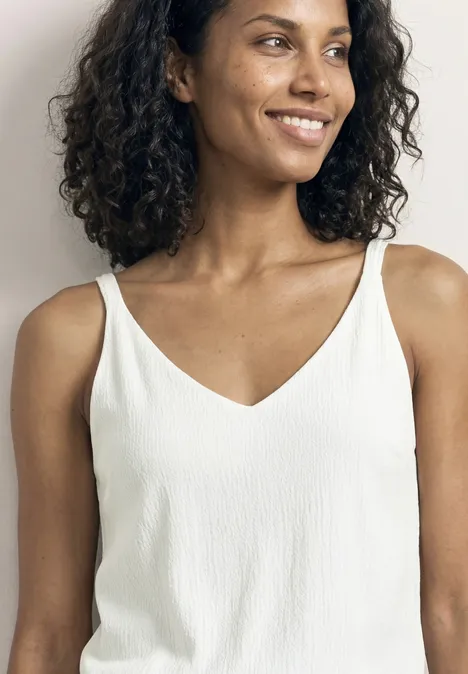 Ein Mensch in weißem ärmellosen Top, lockige Haare, verschwommenes Gesicht.