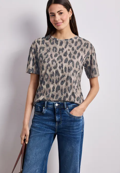 Frau trägt kurzärmeliges Leo-Shirt und blaue Jeans, Hand in Tasche, brauner Trageriemen sichtbar.