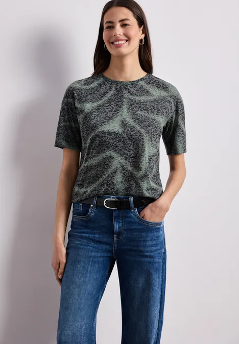 Person in gemustertem Shirt, blauer Jeanshose und Gürtel, posiert vor neutralem Hintergrund.