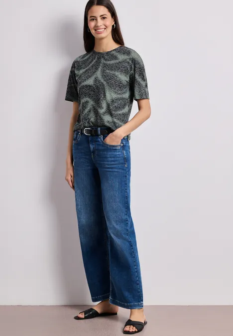 Frau trägt bedrucktes grünes T-Shirt, weite blaue Jeans, schwarzen Gürtel und Sandalen.