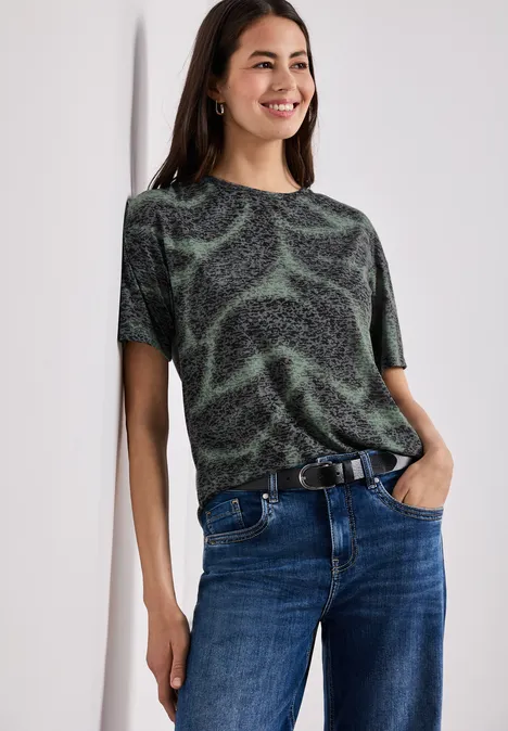 Frau in schwarzem T-Shirt mit grünen Wirbelmustern und Jeans lehnt lässig an der Wand.