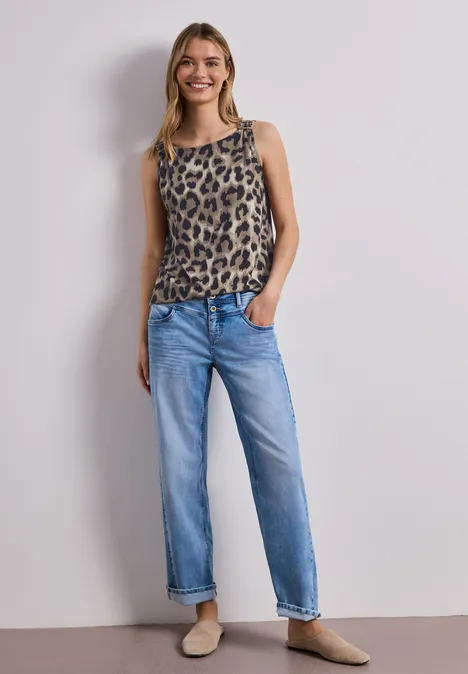 Vrouw voor witte muur, draagt mouwloze top met luipaardprint, blauwe jeans en beige schoenen.