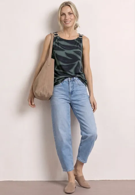 Vrouw in groen-zwart top, lichtblauwe jeans, beige tas en bruine enkellaarzen voor witte achtergrond.