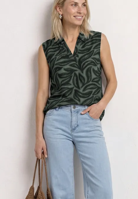 Vrouw in mouwloze donkergroene blouse met patroon en V-hals, lichte jeans en een handtas.
