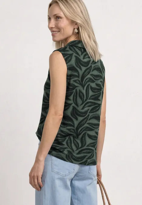 Vrouw van achteren in een mouwloze zwarte top met groene bladprint, lichtblauwe jeans en een lichtbruine tas.