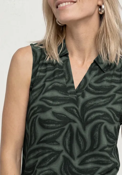 Blonde vrouw in mouwloze donkergroene top met abstract bladpatroon.