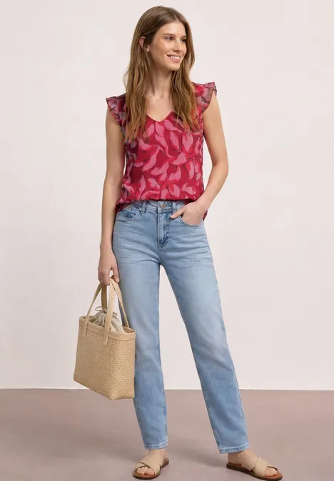Frau trägt pink gemustertes Top, helle Jeans, Sandalen und hält eine Strohtasche vor neutralem Hintergrund.