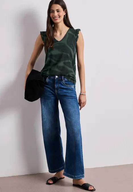 Vrouw voor witte muur met groen mouwloos topje, wijde spijkerbroek, zwarte sandalen en een zwarte tas.