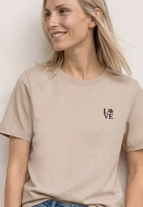 Frau mit beigem T-Shirt und kleinem 'LOVE'-Logo, lächelnd vor hellem Hintergrund.