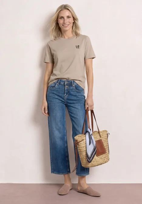 Frau vor hellem Hintergrund mit beigem T-Shirt, weitem Jeans-Culotte, Basttasche und flachen Schuhen.
