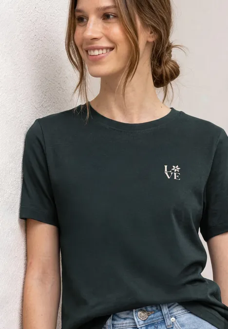 Junge Frau trägt schwarzes T-Shirt mit kleinem weißen „love“-Schriftzug, vor hellem Hintergrund.