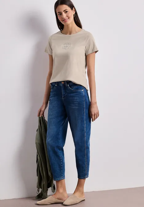 Vrouw draagt beige T-shirt en blauwe jeans, houdt een groene jas vast, casual outfit.