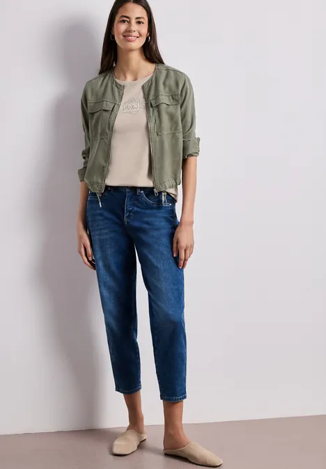 Vrouw in kaki blousejas, beige top, blauwe rechte jeans en beige ballerina’s voor lichte achtergrond.