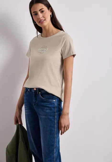 Vrouw met beige T-shirt met klein motief, blauwe jeans en houdt een kaki jas in haar hand.