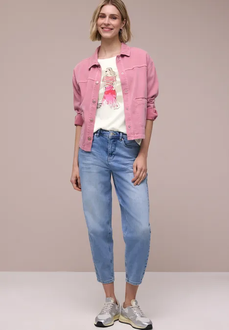 Model draagt roze spijkerjack, wit T-shirt met print, losse blauwe jeans en sneakers tegen neutrale achtergrond.