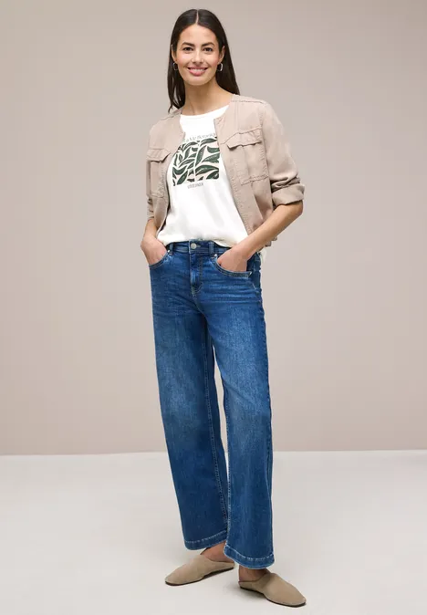 Frau steht vor neutralem Hintergrund, trägt helle Jacke, weißes T-Shirt mit grünem Print, weite Jeans und flache Schuhe.