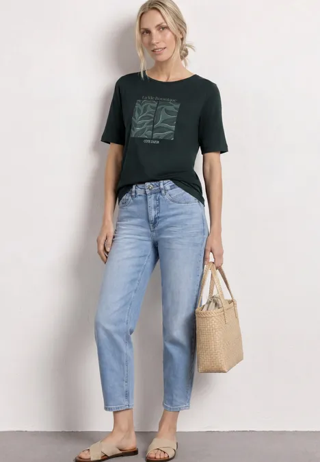 Vrouw in donkergroen T-shirt met print, lichtblauwe jeans en beige tas tegen witte achtergrond.