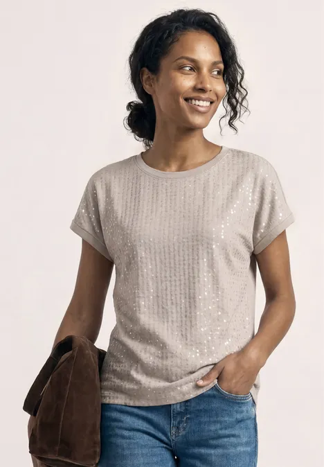 Femme en haut beige à manches courtes à motif discret, jean bleu, tenant une veste marron.