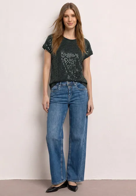 Frau in schwarzem T-Shirt mit Print, blauen geraden Jeans und schwarzen Schuhen vor hellem Hintergrund.