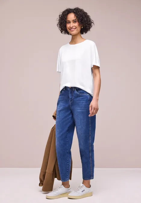 Iemand met wit T-shirt en blauwe spijkerbroek, houdt een bruine jas vast voor lichte achtergrond.