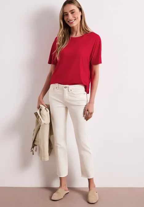 Vrouw in rode T-shirt, witte broek en beige instappers, met een beige jasje in de hand.