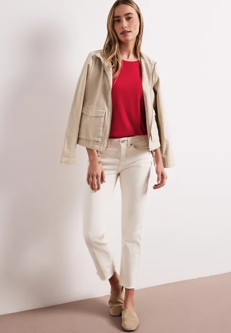 Vrouw in crème jasje, rood topje en lichte broek tegen neutrale achtergrond.