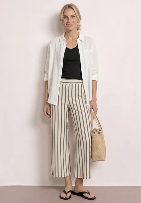 Vrouw met wit hemd over zwart topje, gestreepte wijdvallende broek, slippers en gevlochten tas op neutrale achtergrond.