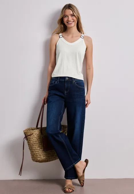 Vrouw in wit topje, donkere jeans, espadrilles en strooien tas voor neutrale achtergrond.