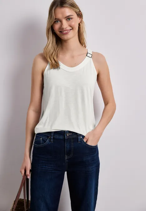 Vrouw draagt witte mouwloze top met gespbandjes en donkerblauwe jeans, met handtas.