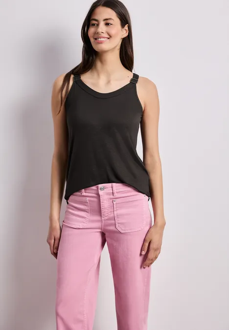 Een vrouw in een zwarte tanktop en roze spijkerbroek staat voor een neutrale achtergrond.