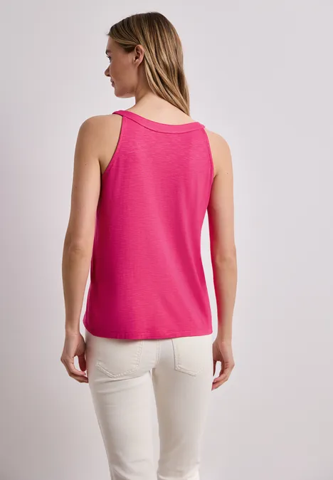 Vrouw van achteren in een felroze mouwloze top en lichte broek tegen neutrale achtergrond.