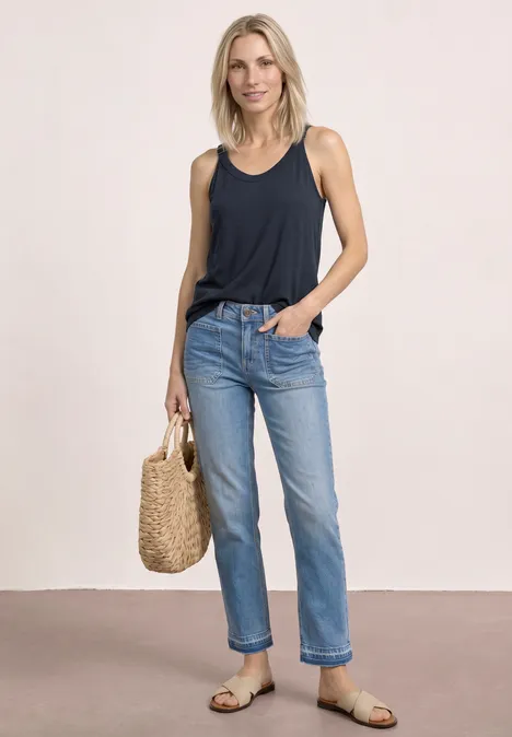 Frau in dunkelblauem Tanktop, hellen Jeans, Sandalen und Strohtasche vor neutralem Hintergrund.