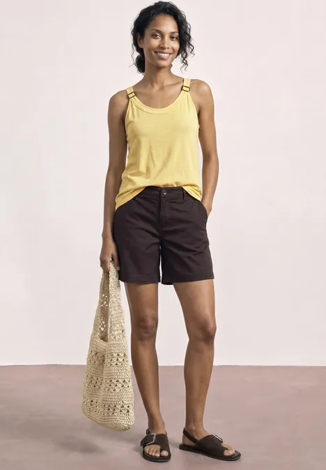 Frau trägt gelbes Top, dunkle Shorts, schwarze Sandalen und eine Netz-Einkaufstasche in der Hand.