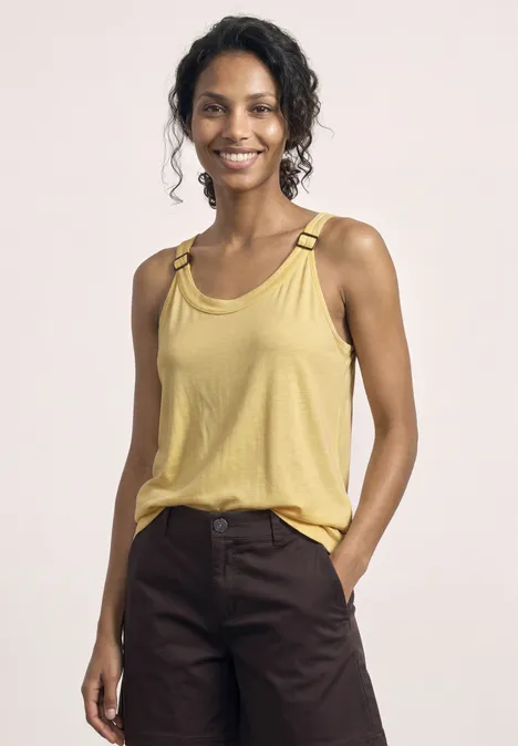 Frau mit lockigem Haar trägt gelbes Tanktop mit Riemendetails und dunkelbraune Hose, eine Hand in der Tasche.
