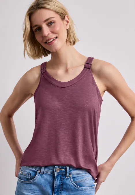 Eine Frau mit hellem Haar trägt ein ärmelloses weinrotes Top und blaue Jeans vor hellem Hintergrund.