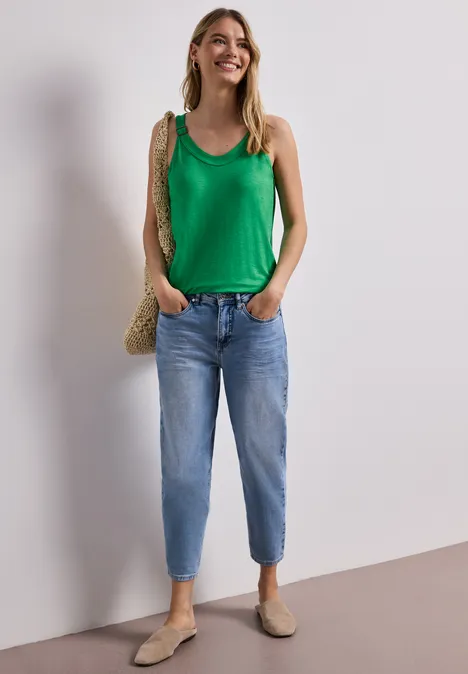 Vrouw voor neutrale achtergrond met groen hemdje, blauwe jeans, beige schoenen en strozak.