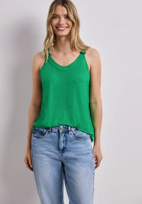 Vrouw draagt groene top en spijkerbroek voor witte muur.