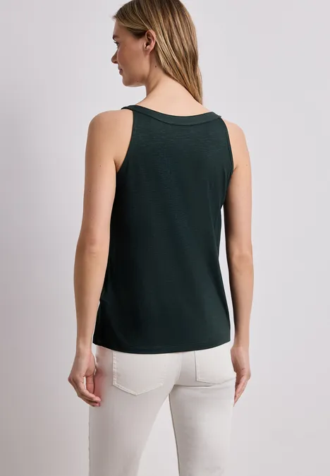 Vrouw van achteren in een groen hemdje en witte broek tegen een neutrale achtergrond.