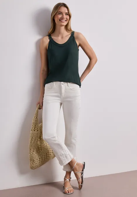 Vrouw in groen hemdje, witte jeans, sandalen en rieten tas leunend tegen een witte muur.