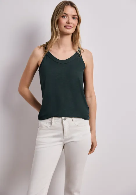 Vrouw in donkergroene top en witte jeans voor een neutrale achtergrond.