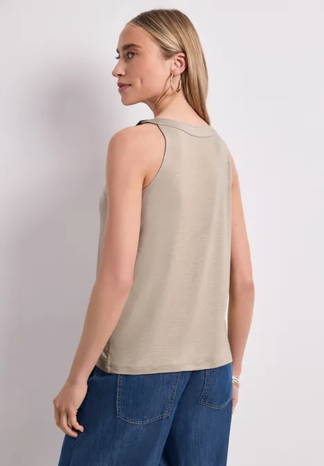 Rückenansicht einer Frau in beigem ärmellosem Top und blauen Jeans vor hellem Hintergrund.