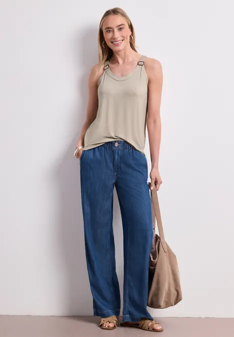 Frau steht in lockerer Pose, trägt beige Top, weite blaue Jeans und eine große beigefarbene Tasche vor weißem Hintergrund.