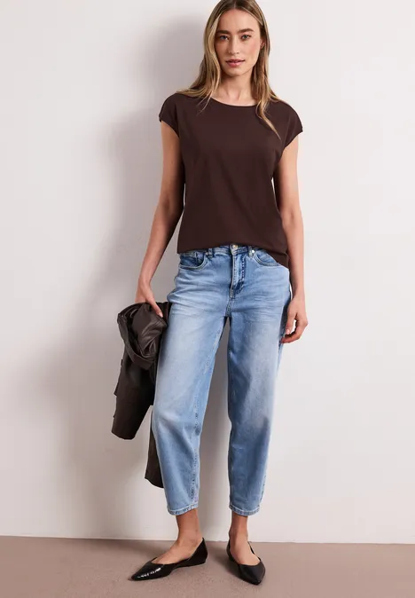 Frau trägt braunes T-Shirt, lockere blaue Jeans, schwarze Ballerinas, hält Jacke in der Hand.