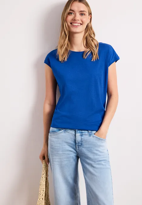 Vrouw in blauw t-shirt en lichtgekleurde spijkerbroek, hand in zak, met strotas tegen witte achtergrond.